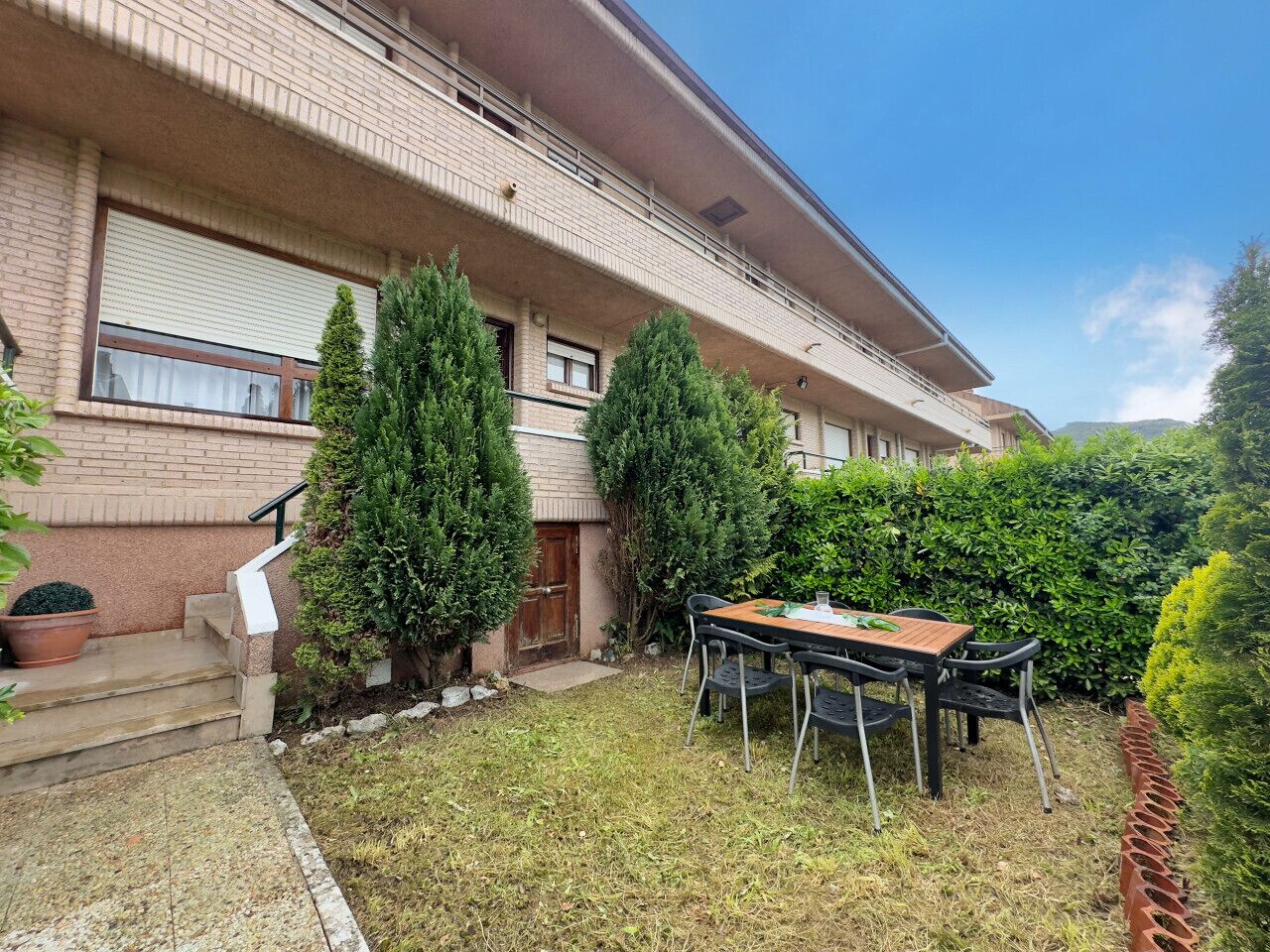 Un edificio residencial con fachada de ladrillo, puerta principal, escalera y un pequeño espacio al aire libre con mesa y sillas.