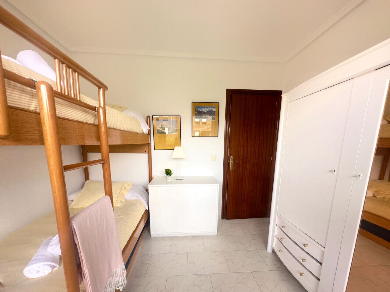 A dormitory room with bunk beds, a desk, a wardrobe, and framed pictures on the wall.