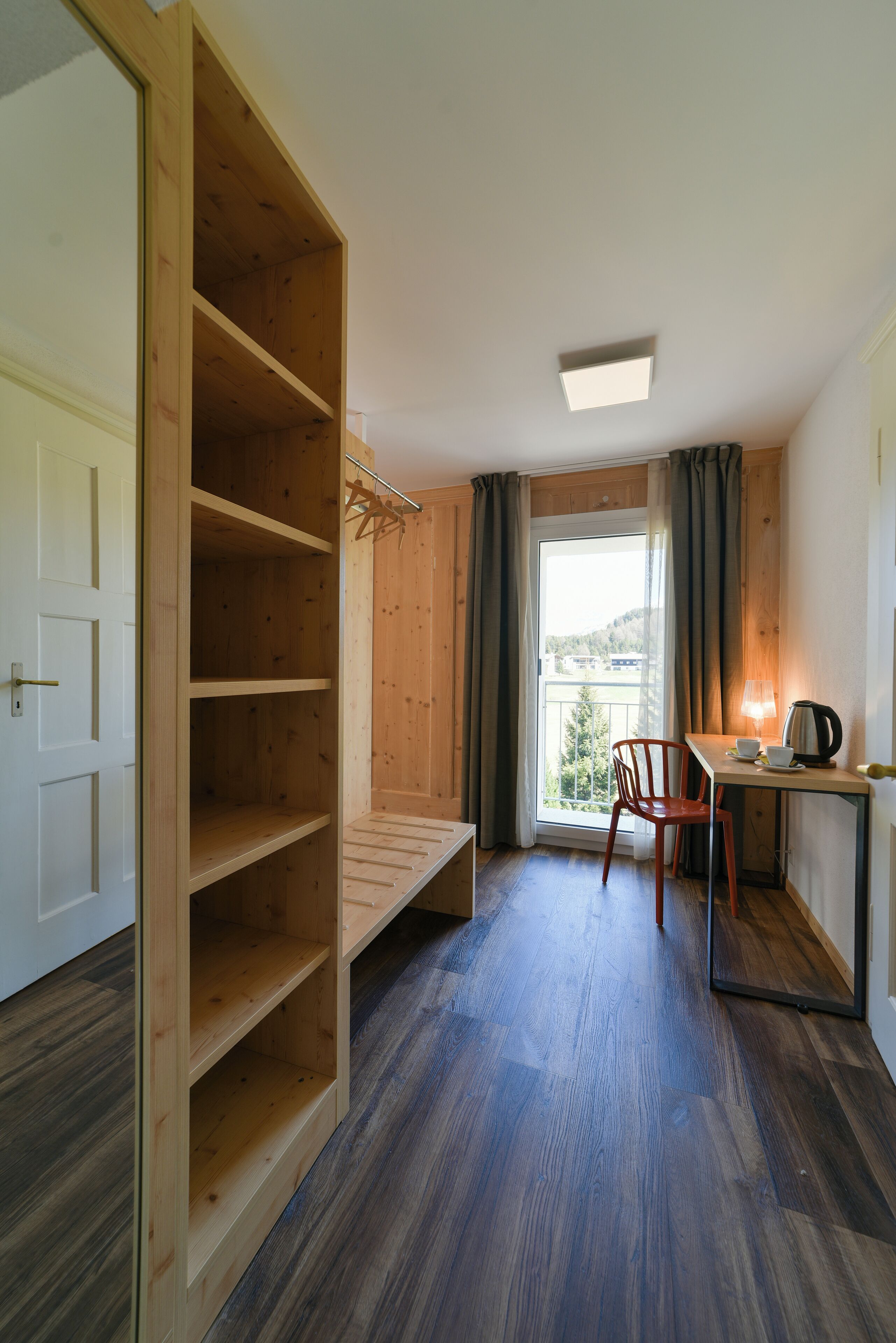 A room with a wooden wardrobe, a desk with a chair, a window with curtains, and a view of the outdoors.