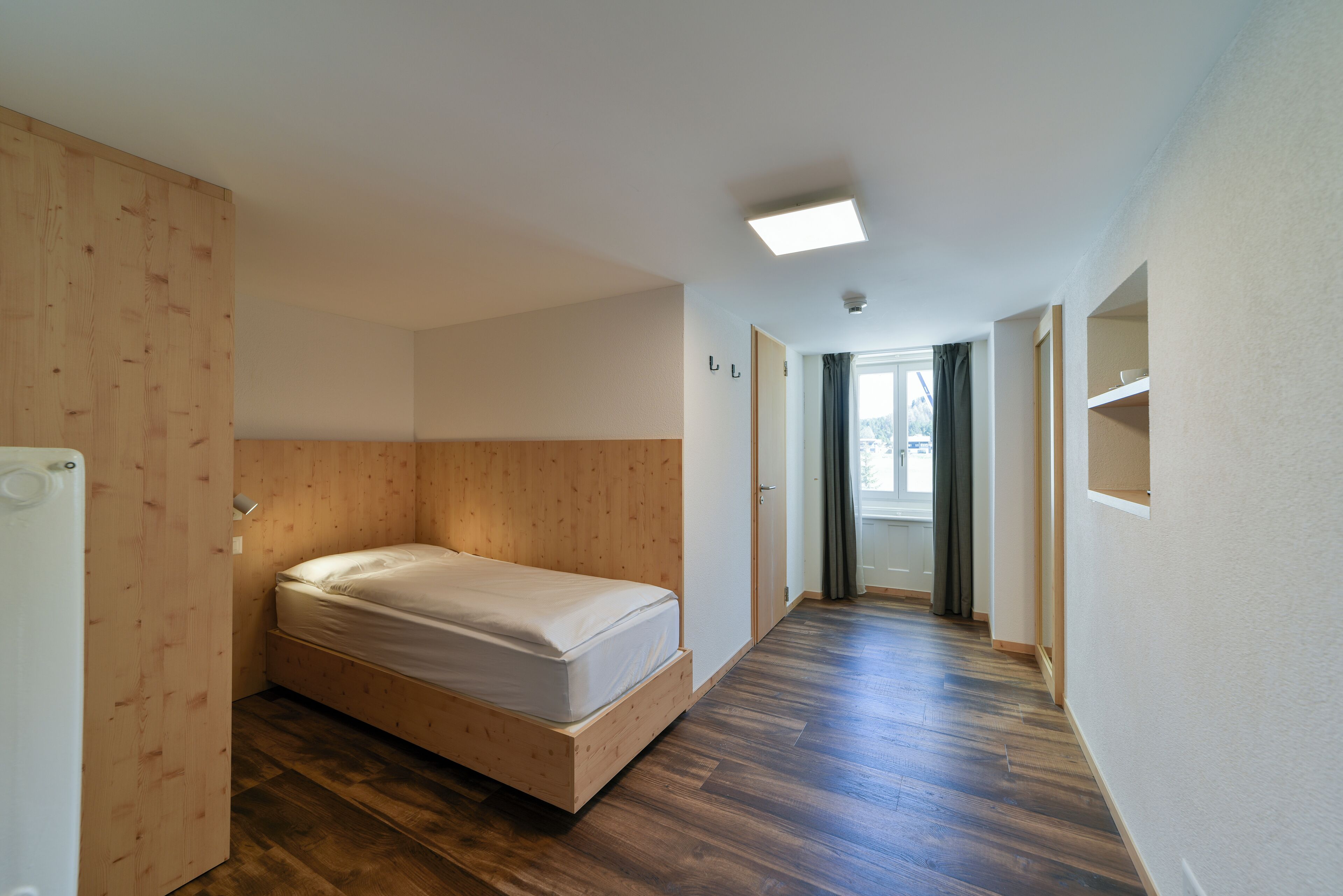 A small, modern room with a wooden bed, a wooden wardrobe, and a long hallway leading to a door with a window.