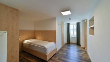 A small, modern room with a wooden bed, a wooden wardrobe, and a long hallway leading to a door with a window.