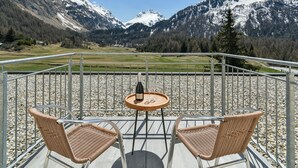 Een balkon met twee rieten stoelen en een kleine ronde tafel, met uitzicht op een bergachtig landschap met besneeuwde toppen en een heldere blauwe lucht.