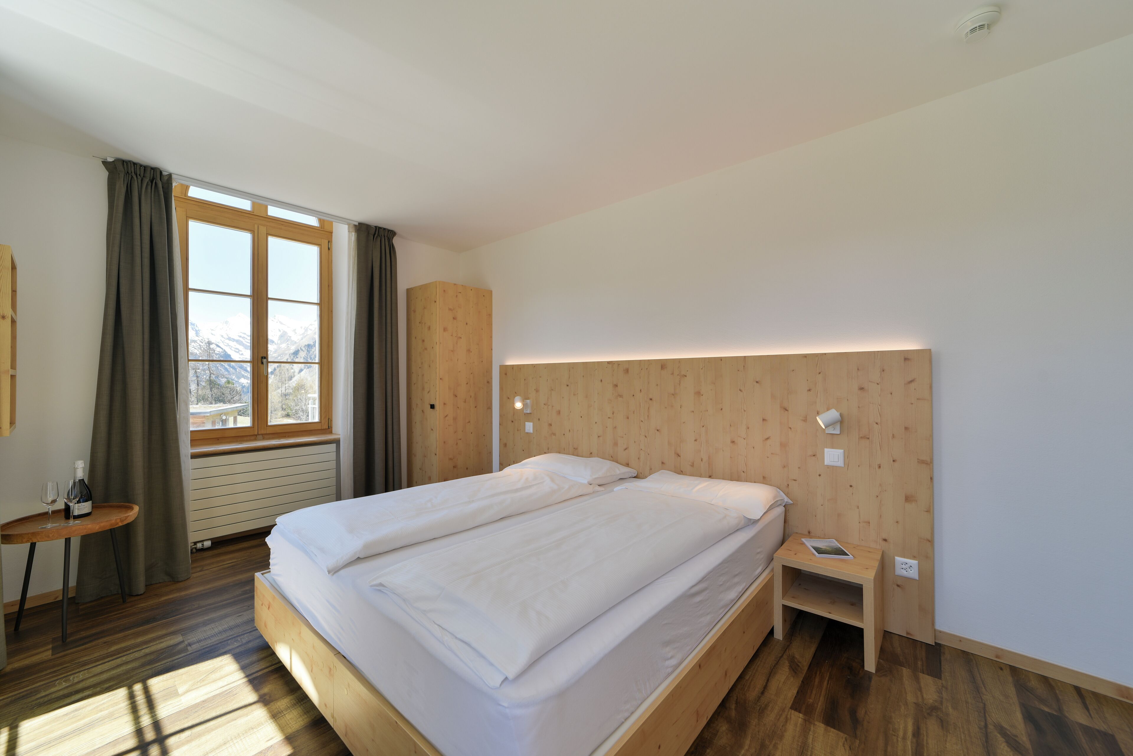 Una habitación de hotel moderna con una cama grande, una mesita de noche con lámpara, una pequeña mesa con una botella y un vaso, y vistas a la montaña desde la ventana.