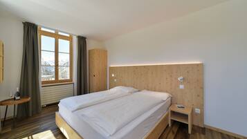 Une chambre d’hôtel moderne avec un grand lit, une table de chevet avec une lampe, une petite table avec une bouteille et un verre, et une vue sur les montagnes par la fenêtre.