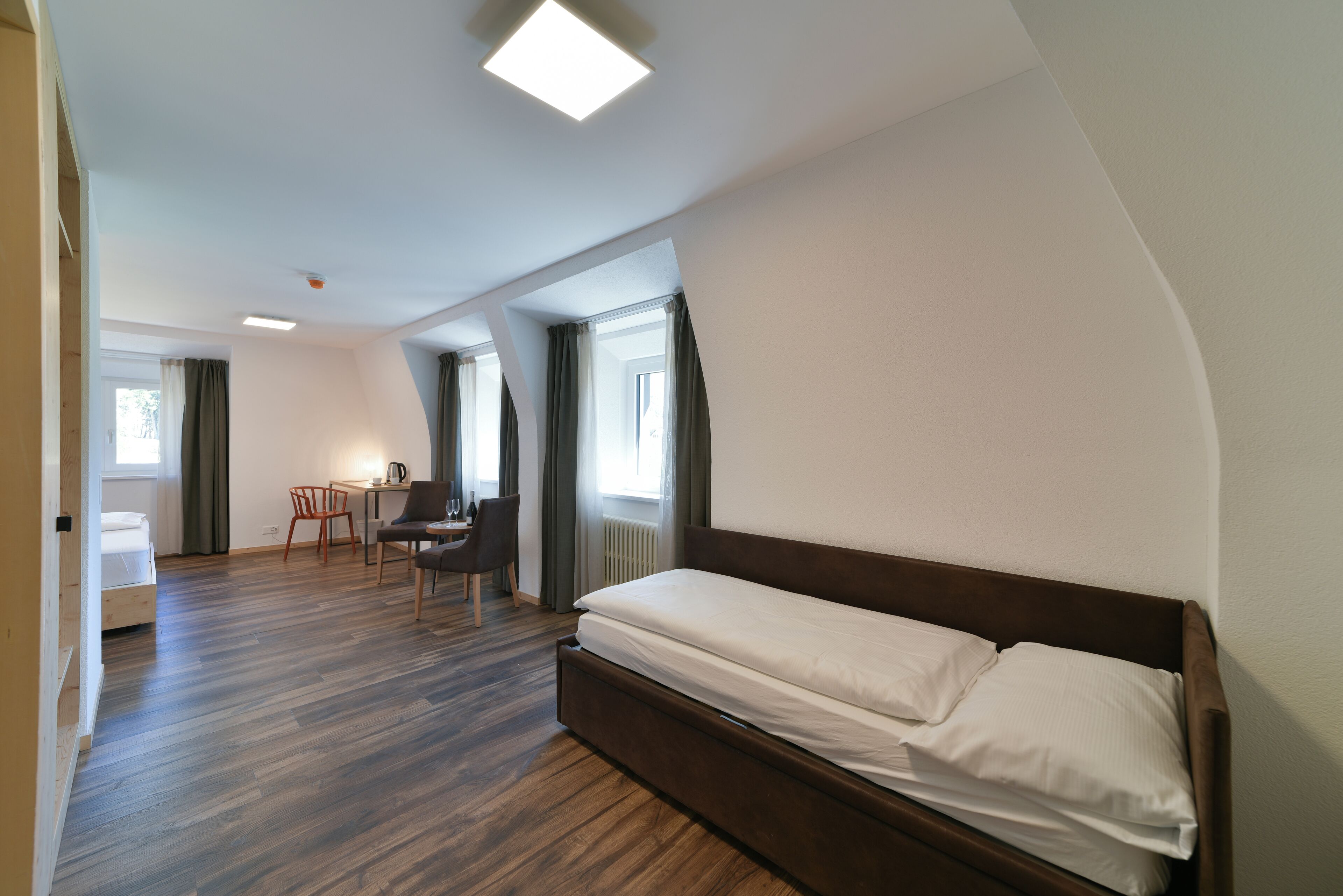 Una habitación de hotel moderna con suelo de madera, una cama individual, un escritorio con una silla y una ventana con cortinas.