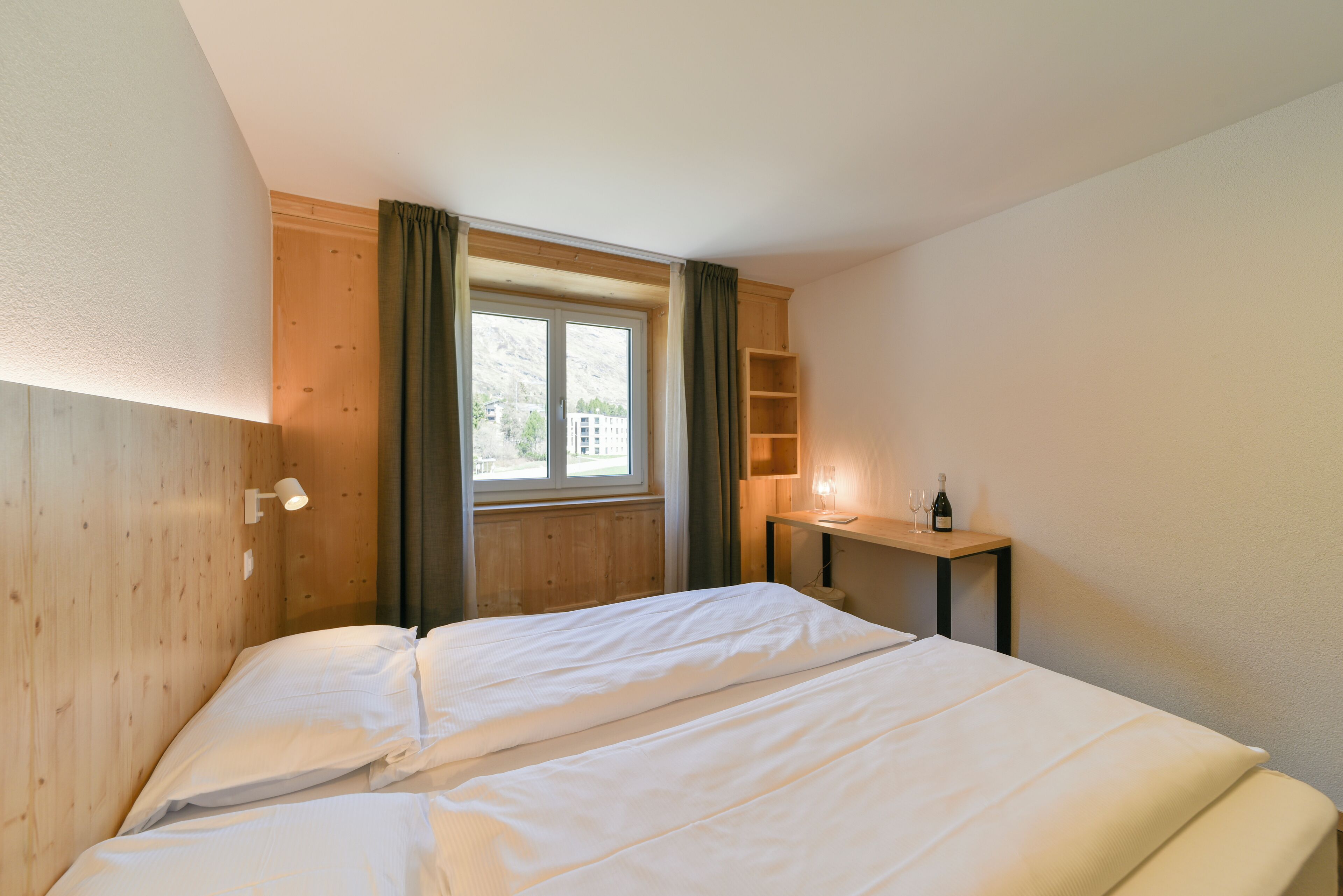 Una cama bien hecha en una habitación con paredes de madera, una ventana con cortinas, una mesita con una botella y vasos, y una estantería.