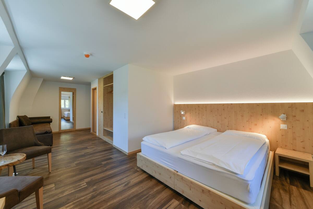 Une chambre d’hôtel moderne avec un plancher en bois, un grand lit avec du linge de lit blanc, une tête de lit en bois, une petite table avec un verre et un canapé.