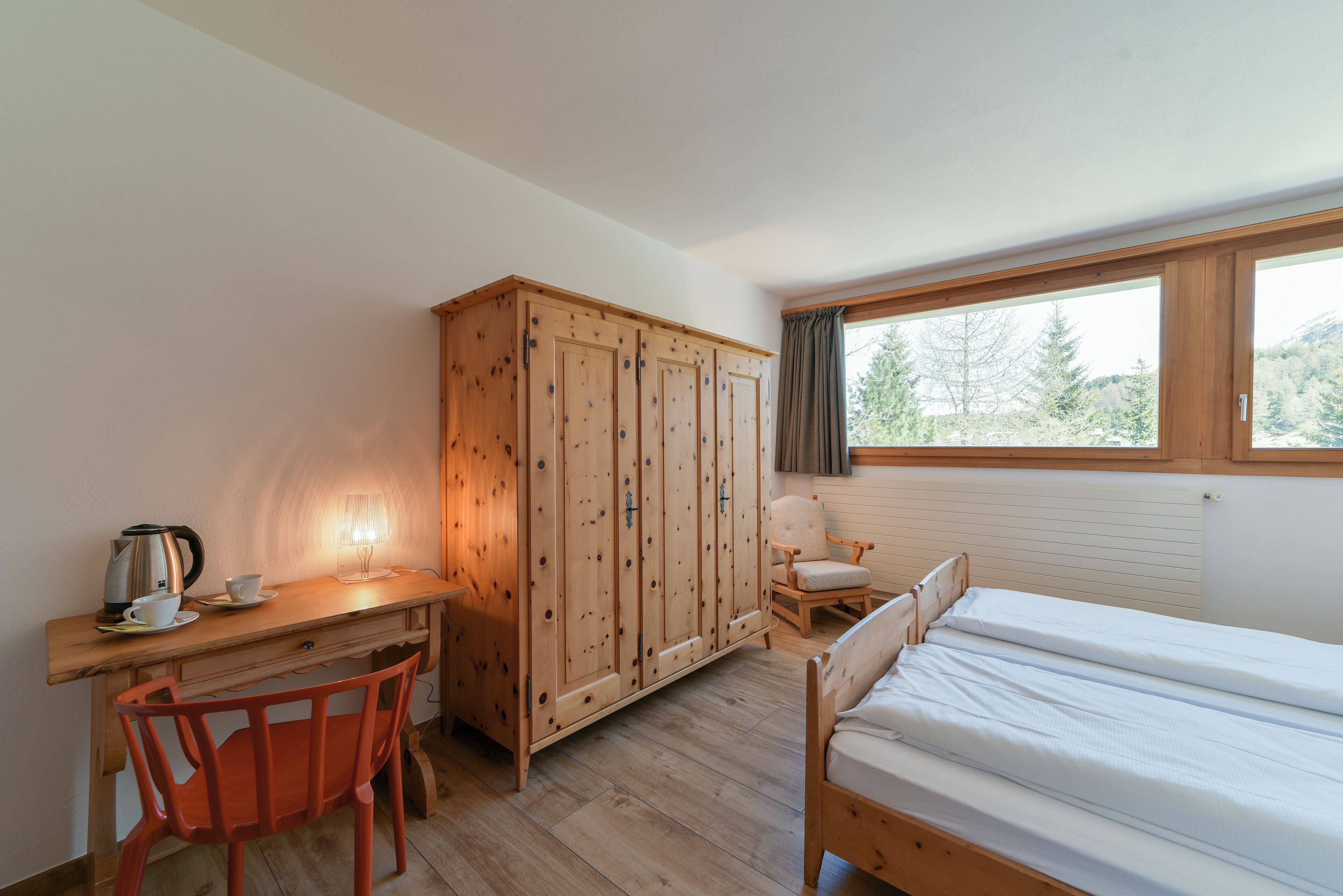 A bedroom with a wooden wardrobe, a desk with a chair, a bed, and a window with a view of trees.