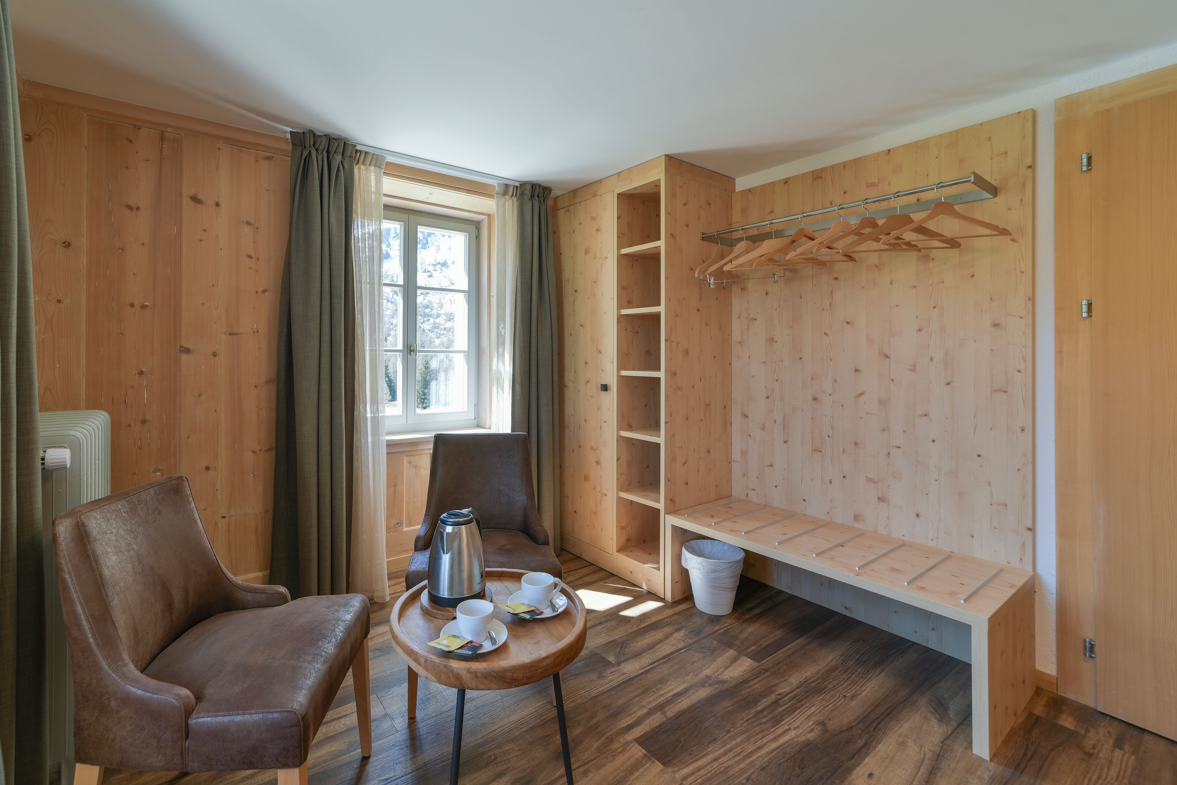 Une pièce aux murs en bois, avec un fauteuil en cuir marron, une petite table sur laquelle se trouvent des tasses et une cafetière, un banc et une armoire en bois dotée d’une tringle pour suspendre des vêtements.