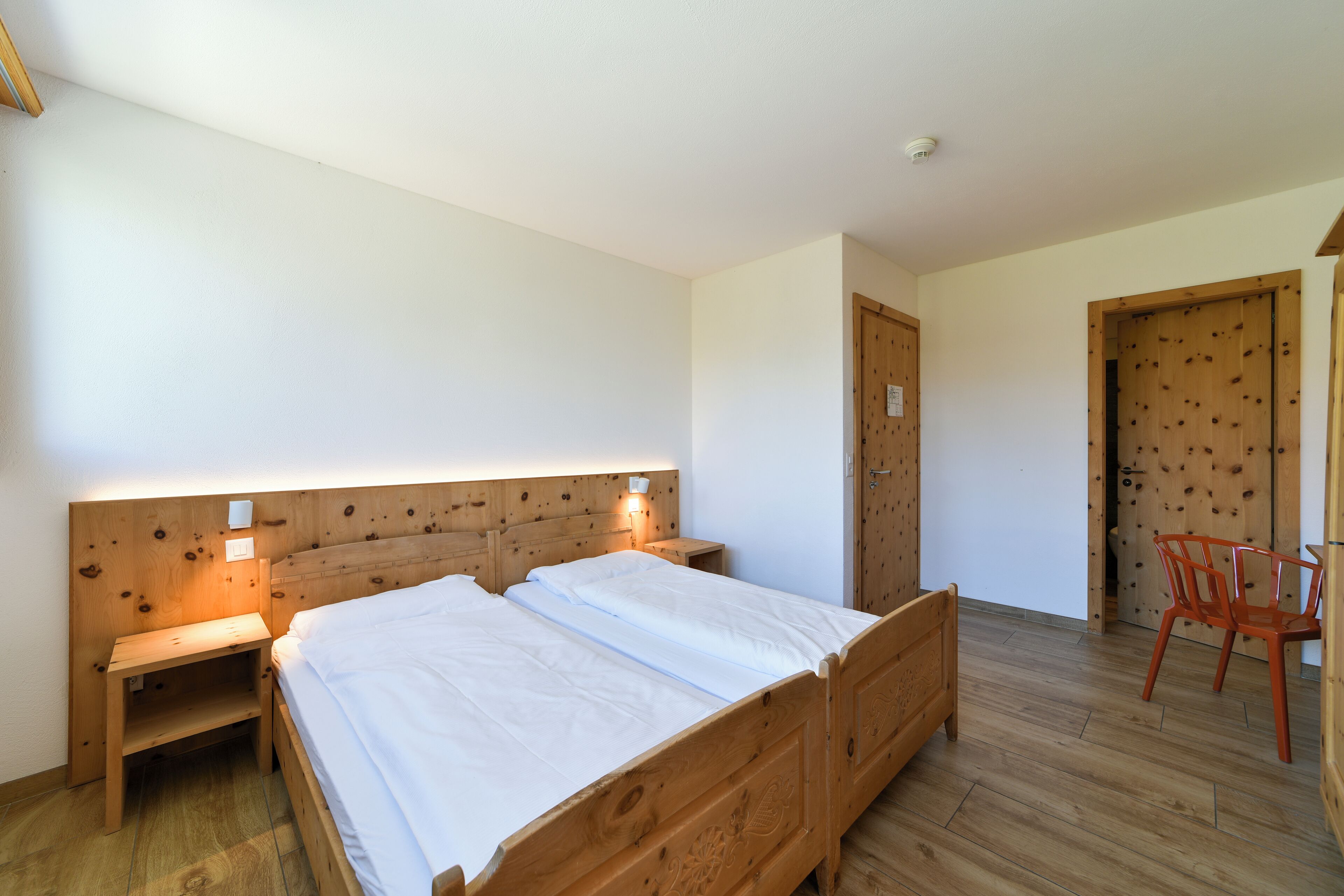 Una habitación con una cama de madera, mesitas de noche, una silla y una puerta.