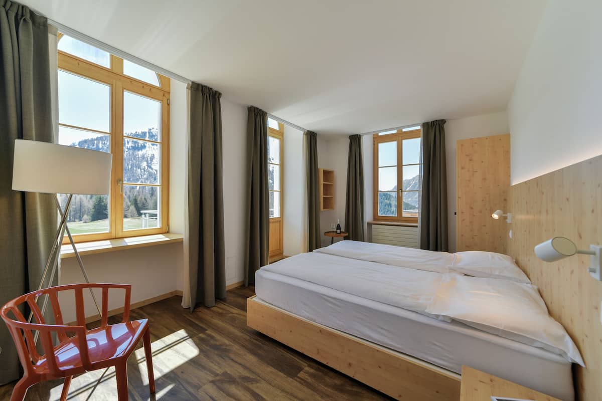 Une chambre avec un lit en bois, une chaise, un bureau et de grandes fenêtres donnant sur une vue imprenable sur les montagnes.