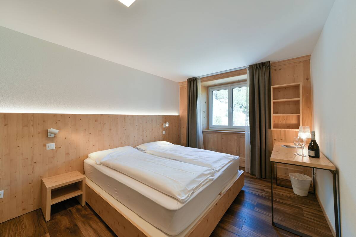 Une chambre à coucher avec un lit en bois, des tables de chevet, un petit bureau avec une bouteille et des verres, et une fenêtre avec des rideaux.