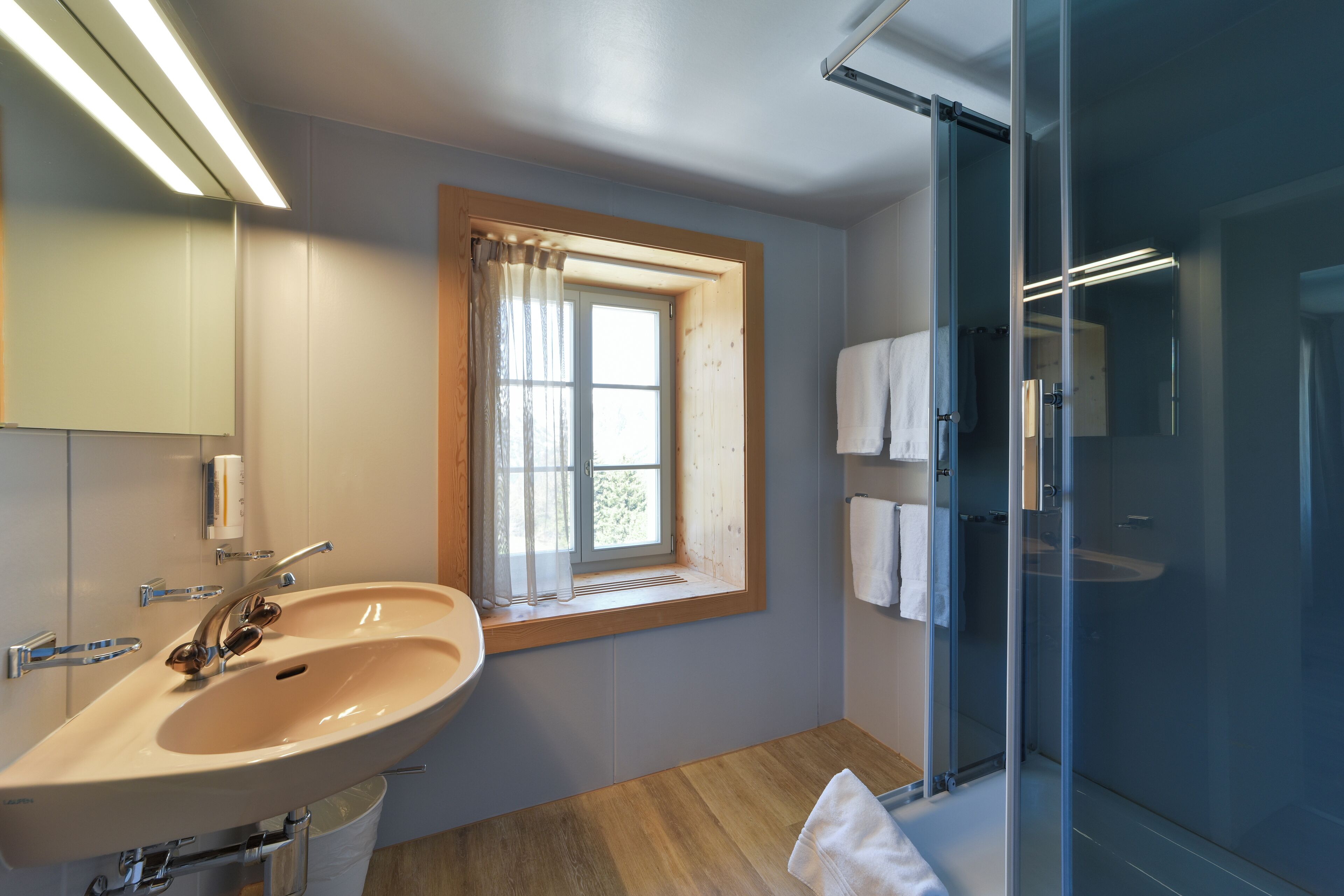 A modern bathroom with a large sink, a window with curtains, a glass shower enclosure, and a towel rack.