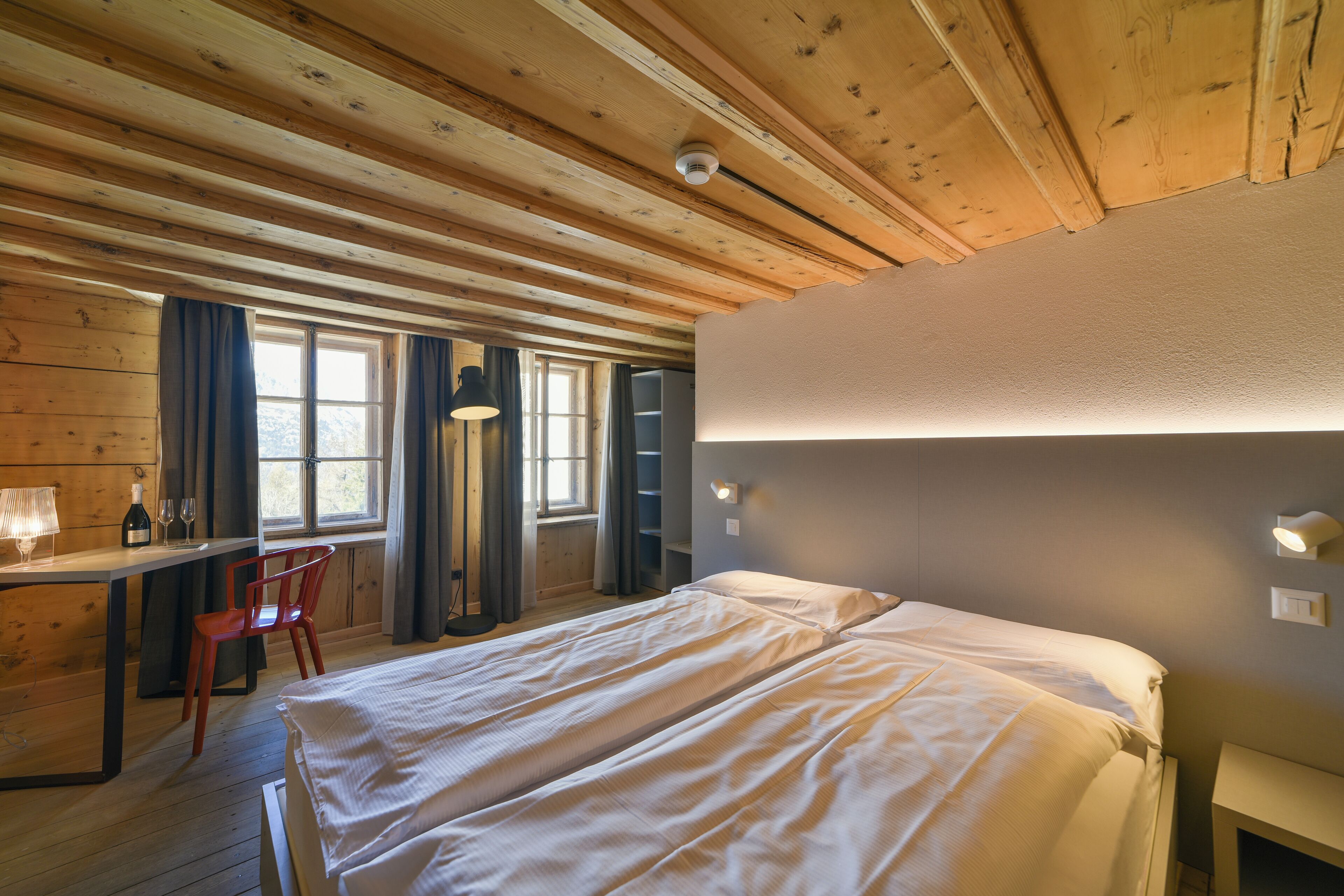 A wooden cabin room with a bed, desk, chair, and a window with curtains.