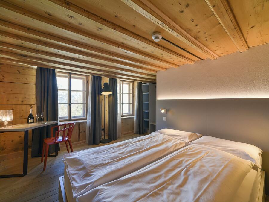 A wooden cabin room with a bed, desk, chair, and a window with curtains.