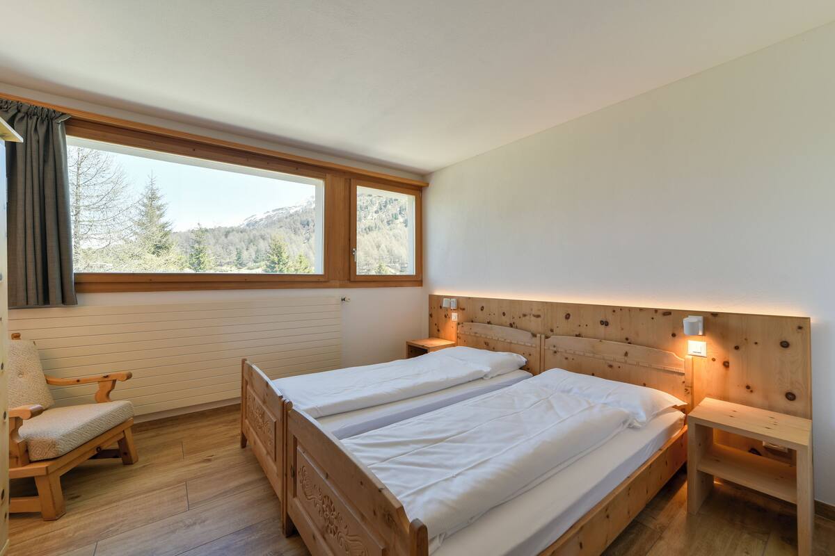 Une chambre avec deux lits simples, une tête de lit en bois, des lampes de chevet, une chaise en bois et une fenêtre donnant sur des arbres et des montagnes.