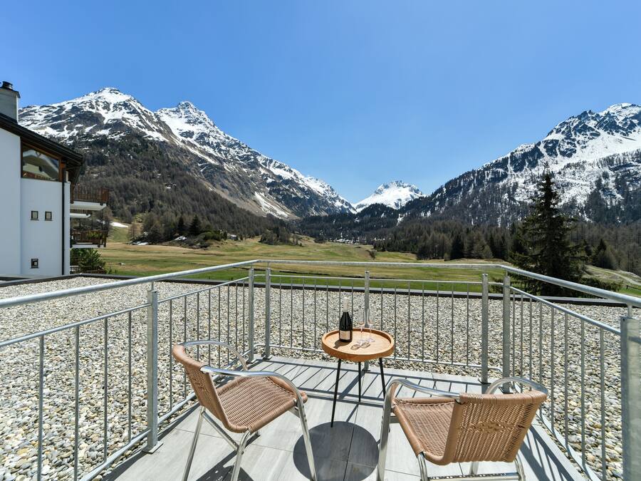 A terrace with wicker chairs and a table, overlooking a mountainous landscape.