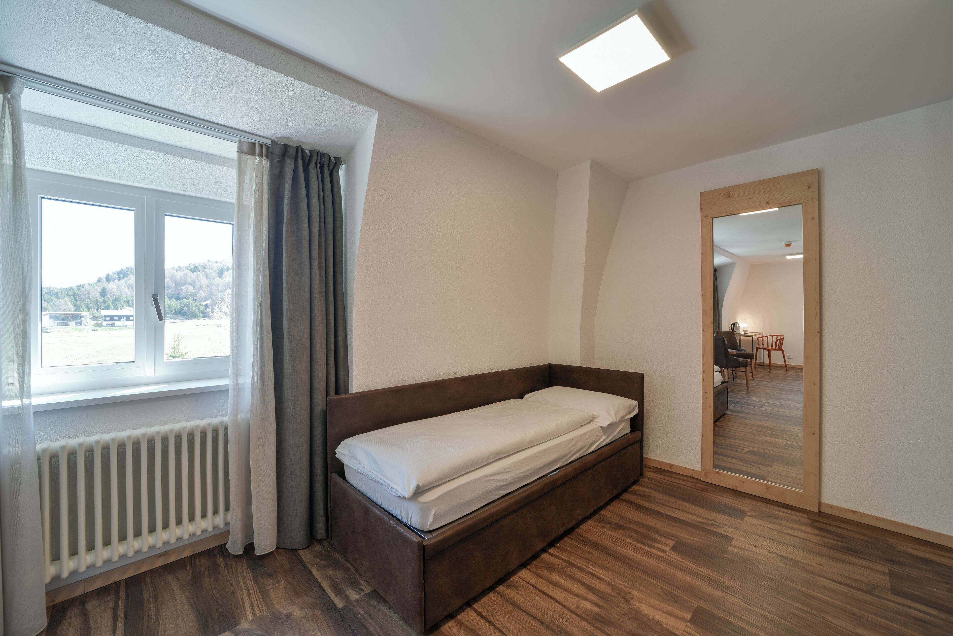 Een moderne slaapkamer met een houten bed, een groot raam met gordijnen, een radiator en een spiegel die de kamer weerspiegelt.