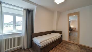 A modern bedroom with a wooden bed, a large window with curtains, a radiator, and a mirror reflecting the room.