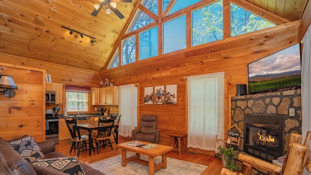 A cozy cabin interior with a stone fireplace, wooden furniture, and a large window overlooking a scenic landscape. - SkiView Hilltop: Honeymoon, Hot Tub, Wifi, Wooded View, Great Location! (Sevierville)