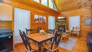 A wooden cabin interior with a dining table, chairs, a fireplace, and a stone wall. - SkiView Hilltop: Honeymoon, Hot Tub, Wifi, Wooded View, Great Location! (Sevierville)