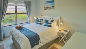 A neatly made bed with a blue throw blanket and rolled towels, a bedside table with a lamp, and a window with curtains. - Francolin's Nest, Langebaan, 4-sleeper (Langebaan)