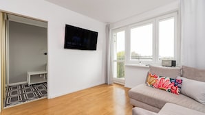 A modern living room with a flat-screen TV, a sofa with colorful cushions, and a view of the outdoors through large windows. - Z widokiem na sopot by Grand Apartments (Sopot)
