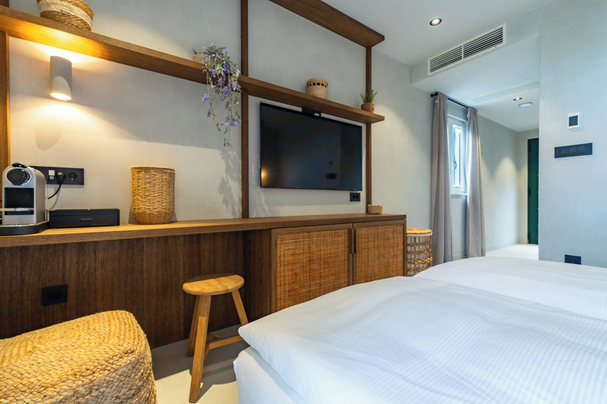 Une chambre d’hôtel moderne avec un grand lit, un bureau en bois doté d’un téléviseur à écran plat, une chaise en osier et un petit tabouret.