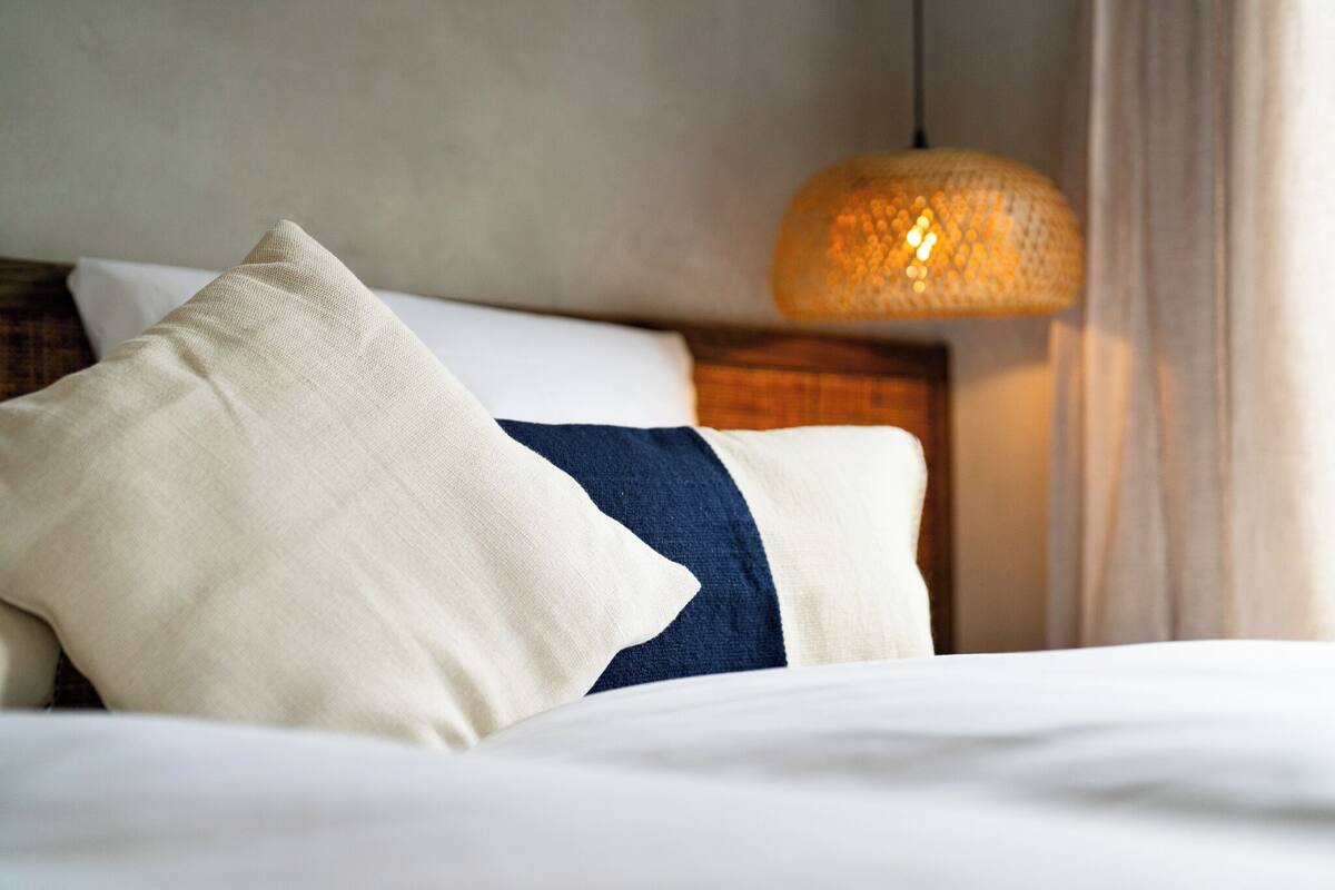 A neatly made bed with white and blue bedding, a woven pendant light, and a wooden headboard.