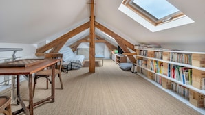 Acogedor dormitorio en ático con techo inclinado, vigas de madera, tragaluces, un escritorio de madera, una silla, una cama y una estantería llena de libros.