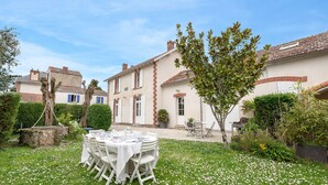Un jardín con una mesa de comedor dispuesta para una comida al aire libre, rodeada de sillas blancas, y un edificio al fondo.