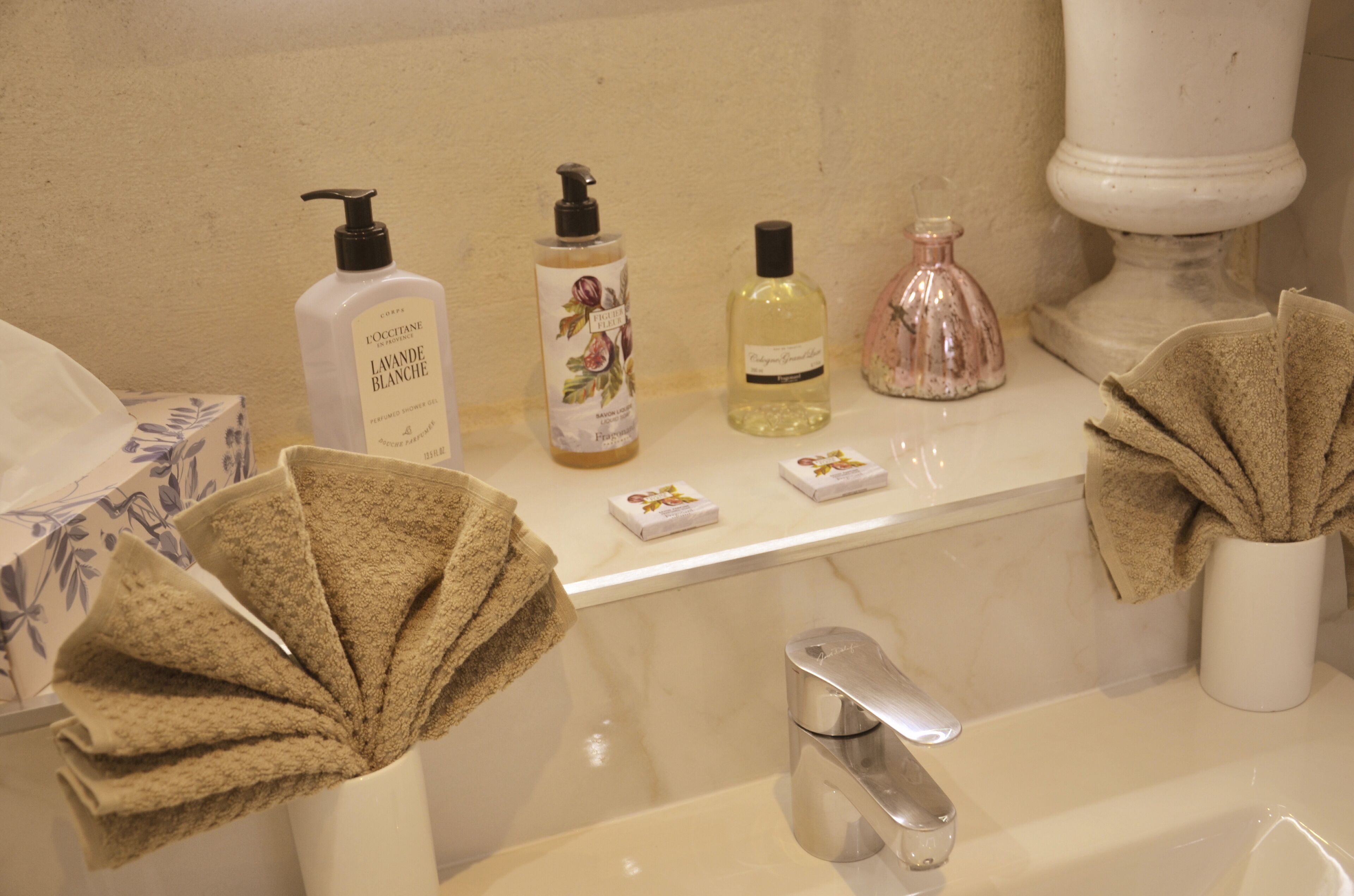 Une salle de bain avec un lavabo blanc, un miroir et des articles de toilette, notamment un flacon de lotion, un distributeur de savon et un flacon de parfum.