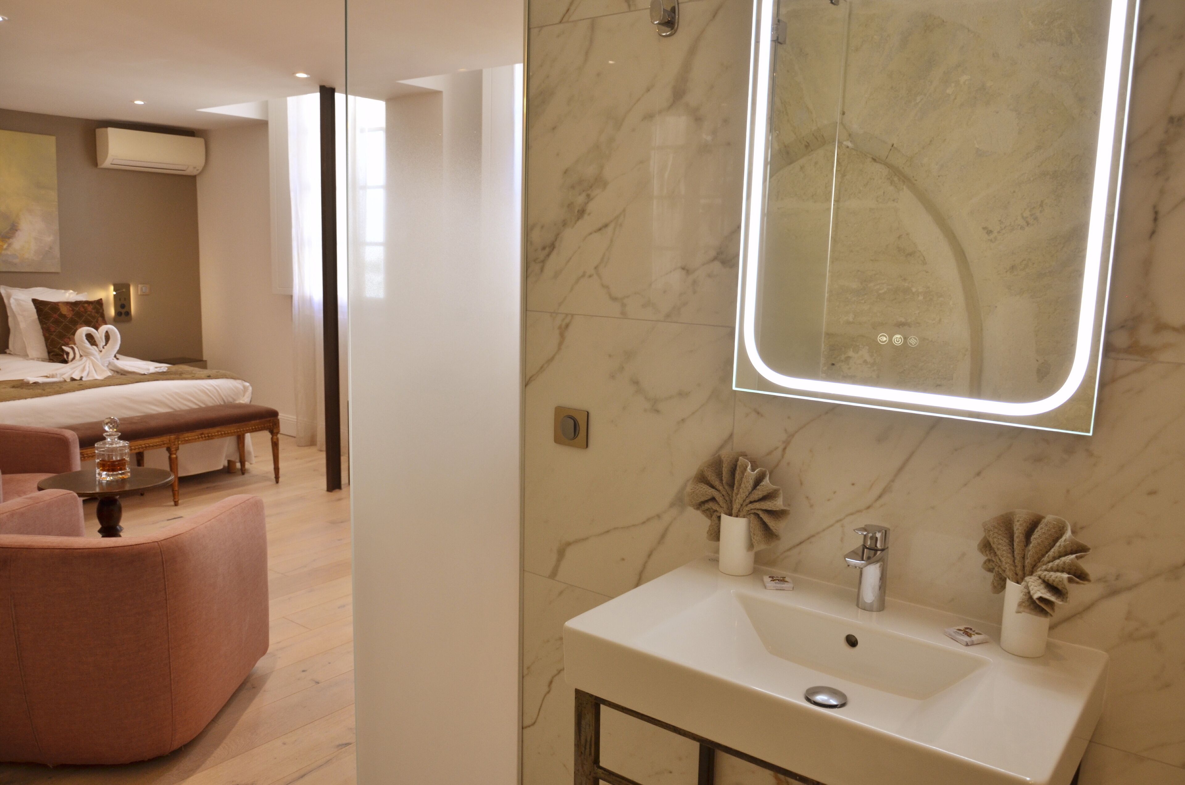 Habitación de hotel moderna con una cama grande, un área de descanso con una silla rosa, un baño de mármol con un espejo amplio y un lavabo blanco.