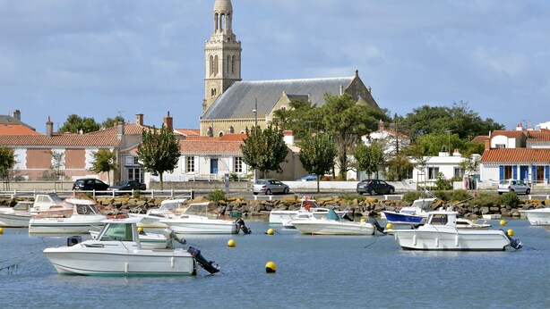 Un port de plaisance où sont amarrés plusieurs bateaux, une église dotée d’un clocher et des bâtiments aux toits recouverts de tuiles rouges.