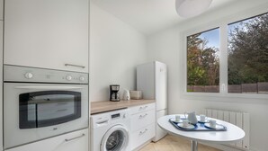 Una cucina moderna con forno integrato, microonde e lavatrice. Si vede una finestra con vista sugli alberi e un tavolo da pranzo bianco con tazze e un vassoio.