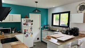 A modern kitchen with a teal accent wall, white cabinetry, a built-in island with stools, a coffee machine, and a window with a view. - 3-bedroom house with garden in Arès, 500m from beach and lake (Arès)