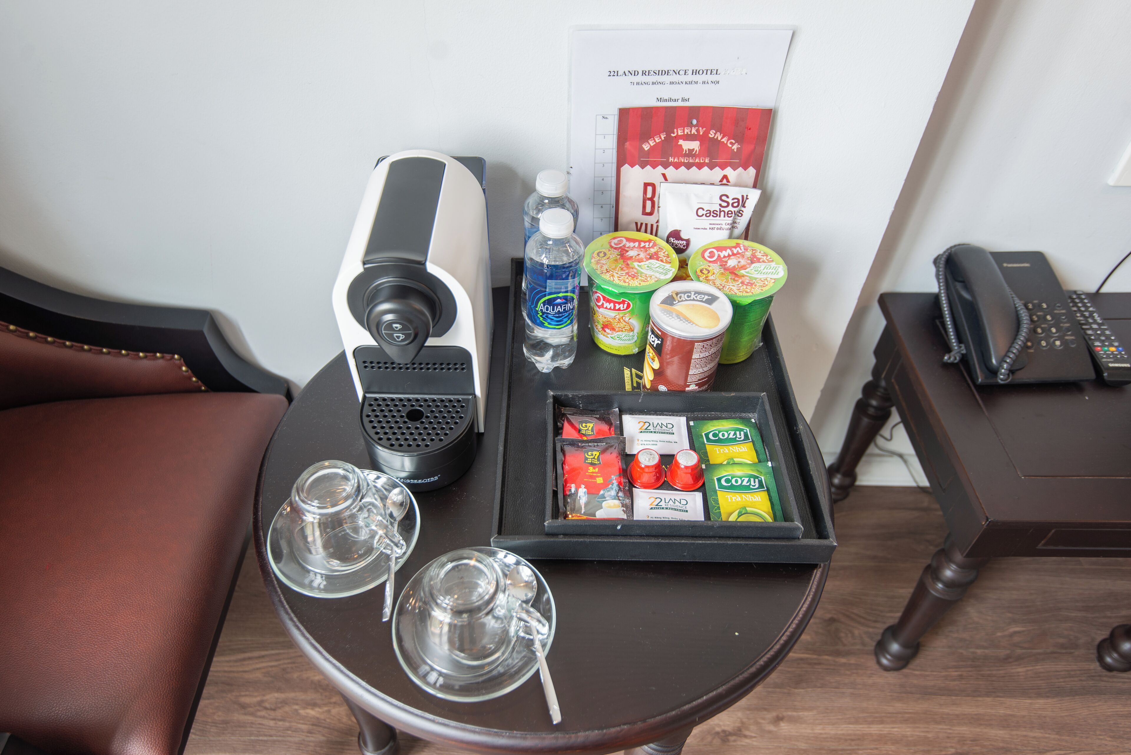 A hotel room with a small table containing a coffee maker, water bottles, snacks, and a phone.