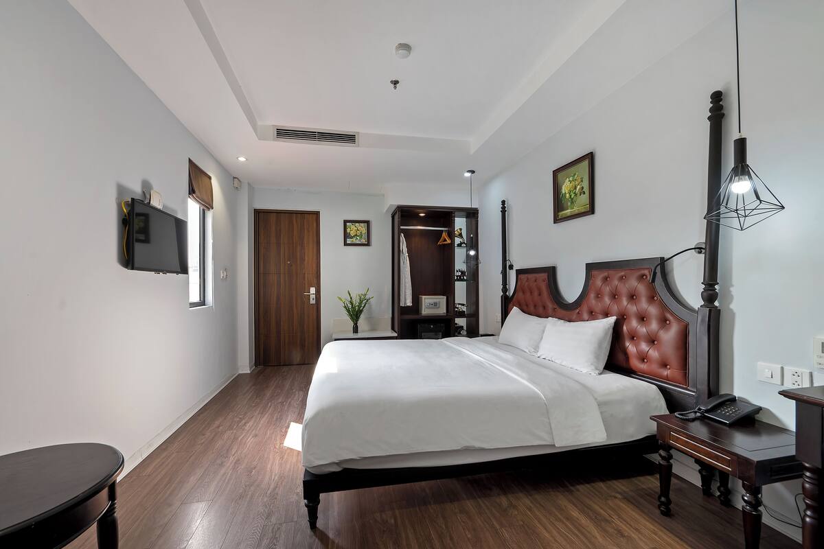 A neatly arranged bedroom with a large bed, a bedside table with a lamp, a TV mounted on the wall, and a wooden door.