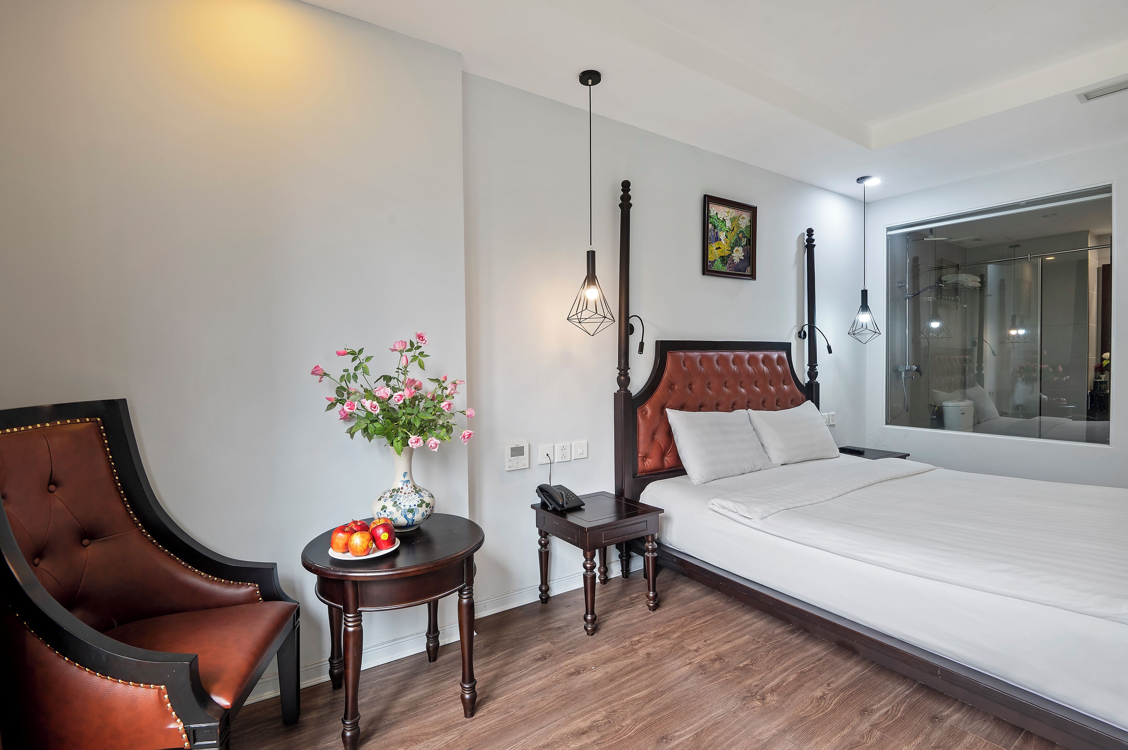 A hotel room with a large bed, a wooden chair, a small table with a vase of flowers, and a nightstand with a phone.
