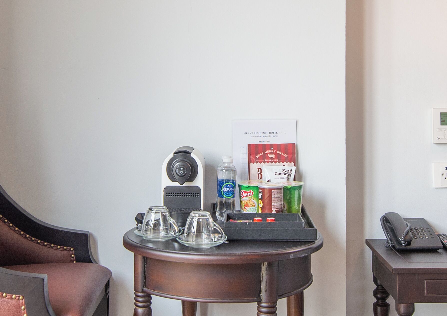 A small wooden table with a coffee maker, glasses, a water bottle, and a snack package in a hotel room.