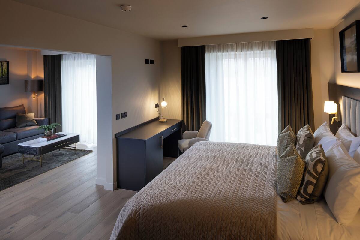 Ein modernes Hotelzimmer mit einem großen Bett, einem Schreibtisch, einer Couch und Blick ins Freie.