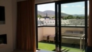 A modern living room with a sofa, a coffee table, and a view of a balcony with outdoor furniture. - House 15 (Kenmare)