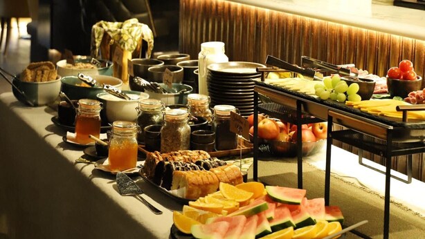 A buffet table with various food items and condiments, a bar area with glasses and bottles, and a brick wall with a patterned design in the background.