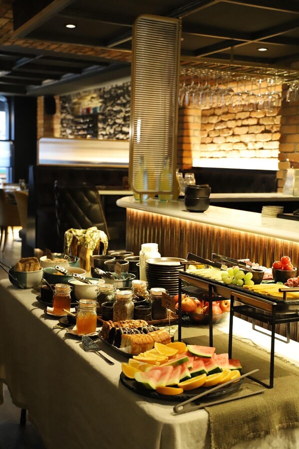 A buffet table with various food items and condiments, a bar area with glasses and bottles, and a brick wall with a patterned design in the background.