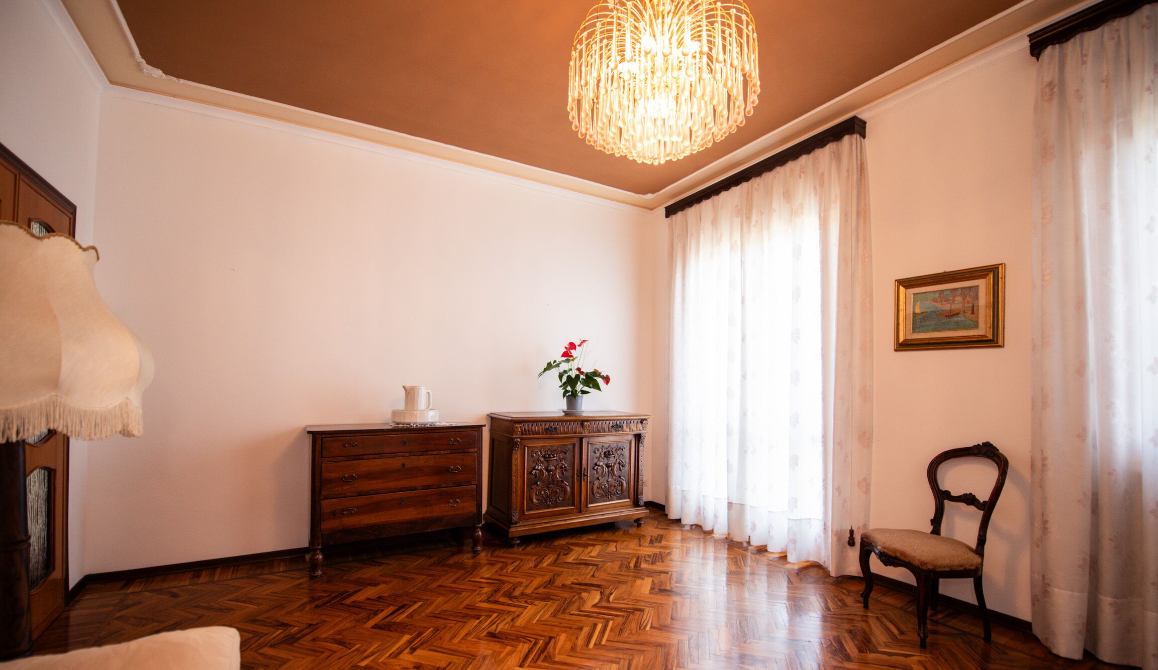 Um quarto com piso de madeira em padrão herringbone, um armário de madeira, uma cadeira, uma mesa com um vaso de flores, um candelabro e um quadro na parede.