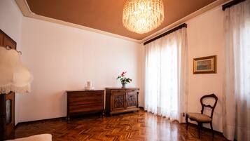 Um quarto com piso de madeira em padrão herringbone, um armário de madeira, uma cadeira, uma mesa com um vaso de flores, um candelabro e um quadro na parede.