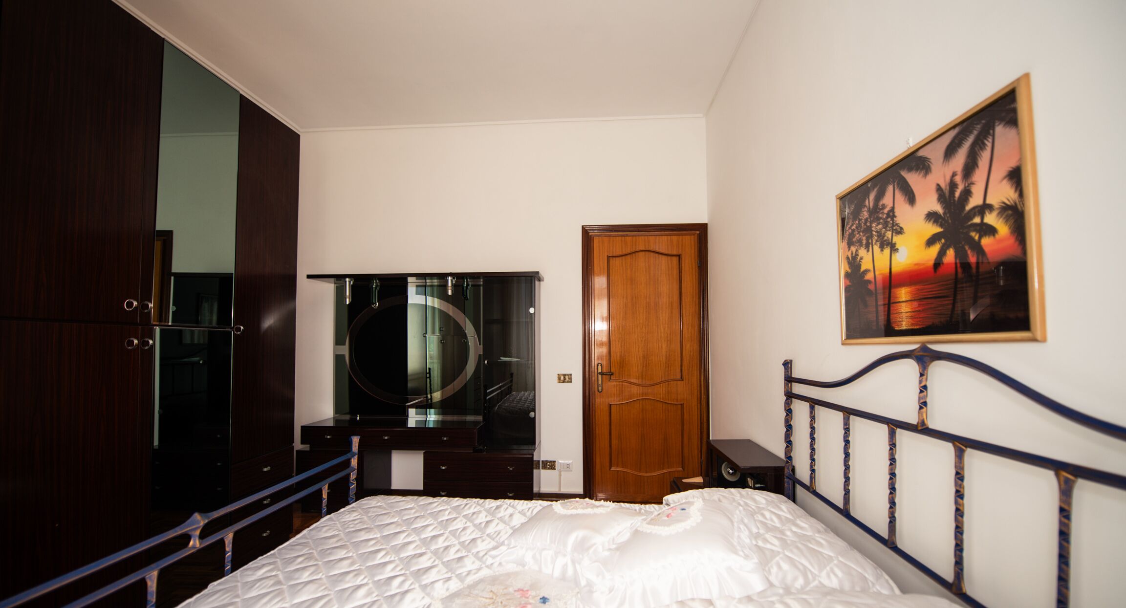 Ein Schlafzimmer mit einem Bett, einem Kleiderschrank, einem Spiegel und einem Sonnenuntergangsbild.