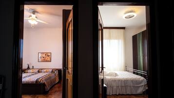 Dois quartos com piso de madeira, um com colcha estampada e um ventilador de teto, e outro com colcha lisa e uma mesa de cabeceira.