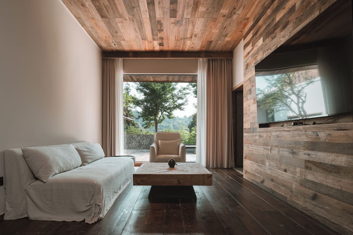 Un salon moderne avec un plafond en bois, un canapé, une table basse et une grande fenêtre donnant sur la forêt.