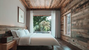 Un dormitorio con techo de madera, una cama grande, una mesita de noche, una ventana con persianas y vistas a árboles.